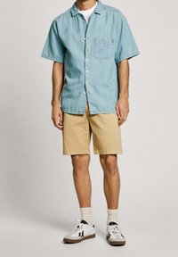Camisa de mezclilla de manga corta azul claro con un bolsillo, combinada con pantalones cortos beige y zapatillas blancas, con detalles en negro y calcetines a rayas.