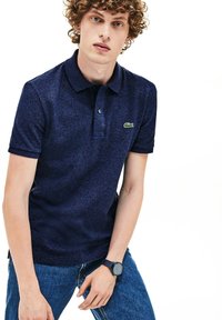 Polo marine à manches courtes en tissu texturé, avec trois boutons et un logo de crocodile vert ; porté avec un jean bleu et une montre.