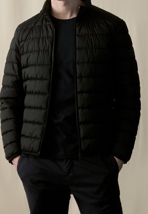 Homme portant une veste matelassée noire zippée par-dessus un t-shirt noir, les mains dans les poches, sur un fond beige uni.