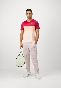 Herreidrettsbekledning med en maroon- og rosa gradient T-skjorte, lyserosa bukser og hvite sneakers, holder en tennisracket.