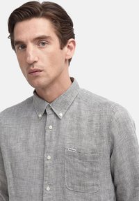 Mann mit kurzen braunen Haaren, der ein knopfgaffiertes graues kariertes Hemd mit einer Brusttasche und Markenschild trägt, schaut auf einen einfarbigen Hintergrund.