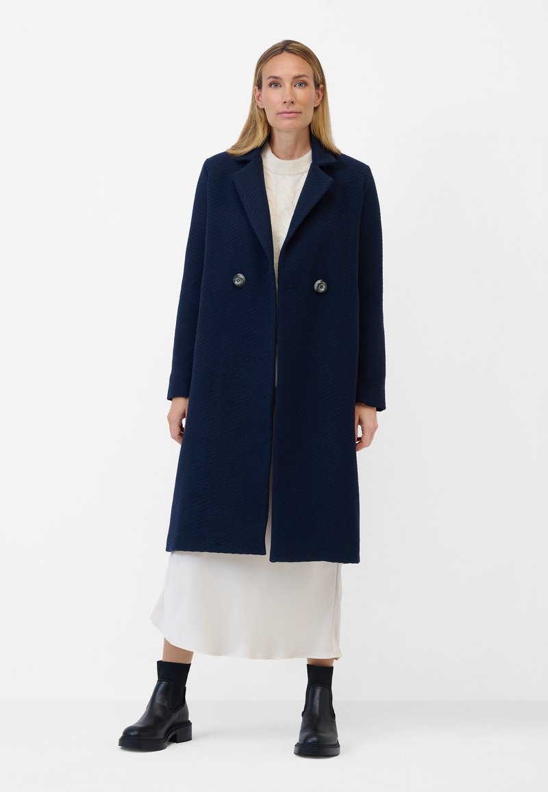 Marinblå dubbelknäppt kappa med texturerad tyg, stora slag och två knappar. Bärs över en vit klänning och matchas med svarta ankelstövlar.