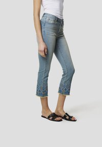 Lichtblauwe cropped jeans met decoratieve blauwe en gouden borduursels bij de zoom, gecombineerd met zwarte sandalen met uitgesneden accenten.