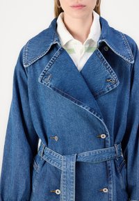 Denim plašč z veliko ovratnikom, dvorednim gumbom in zavezanim pasom. Ima modro barvo z opaznimi šivi.