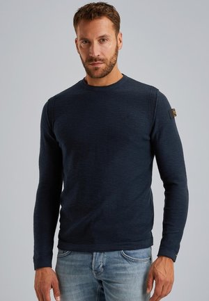 Marineblauer Langarm-Pullover mit gerippten Schulterdetails und kleinem Logo auf der Brust. Der Stoff wirkt weich und gestrickt. Kombiniert mit Jeans.
