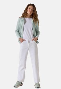 Felpa verde chiaro con zip sopra una maglietta bianca, abbinata a pantaloni bianchi larghi e scarpe da ginnastica verdi con dettagli grigi.