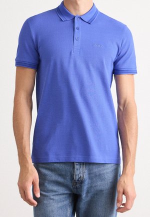 Homme portant un polo bleu cintré avec des boutons et le logo « BOSS », associé à un jean denim bleu clair, sur un fond uni.