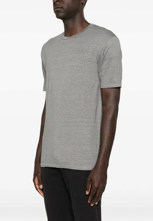 Homme portant un t-shirt uni gris clair à manches courtes et un pantalon noir, debout devant un fond blanc.