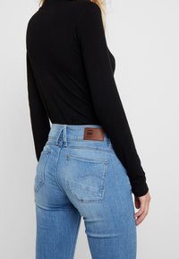 Jeans de mezclilla azul claro con una forma ajustada, que muestran los bolsillos traseros y una etiqueta de cuero marrón. Se puede ver una camiseta negra de manga larga.