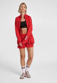 Rotes Sportjacken- und Shorts-Set mit grauen Akzenten. Besteht aus einer Jacke mit Reißverschluss und einem verkürzten schwarzen Tanktop. Enthält gestreifte weiße Socken und Sneakers.