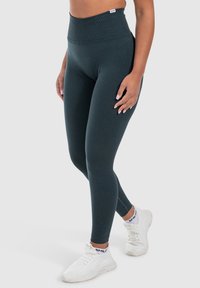 Hoge taille leggings van donkergroene stof; gladde textuur en aansluitend ontwerp. Gedragen met witte sportschoenen.