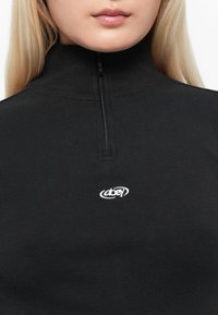 Črna rebrasta zip-up majica z visokim ovratnikom, ki ima majhen bel vezeni logotip "obey" na prsih. Gladka tekstura in oprijet dizajn.