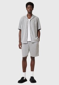 Camicia a maniche corte in affeltrito grigio chiaro con bottoni, abbinata a pantaloni corti coordinati e scarpe slip-on nere con calzini bianchi.