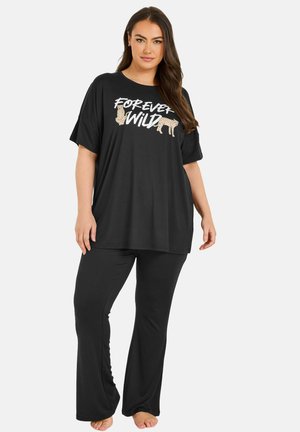 Frau mit langen braunen Haaren, die ein lockeres schwarzes T-Shirt mit der Aufschrift "Forever Wild" und Tierkonturen trägt, kombiniert mit schwarzen Hosen und barfuß.