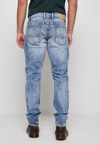 Homme portant un jean bleu clair délavé et des bottes marron foncé, debout dos à un arrière-plan clair uni.