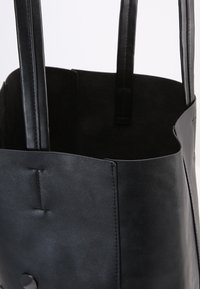 Černá kožená tote taška s hladkou texturou, dvěma plochými uchy, minimalistickým designem a kulatým detailem tlačítka na přední straně.