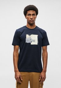 Homem com cabelo afro vestido com uma t-shirt gráfica azul-marinho e calças cargo castanhas, de pé contra um fundo claro e liso.