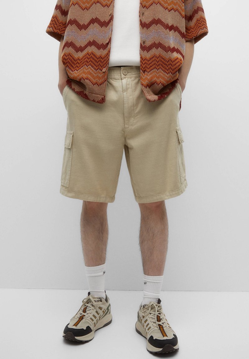 Shorts cargo beiges en coton, avec poches latérales, coupe droite, associés à une chemise en tricot à motif et des chaussures de sport.