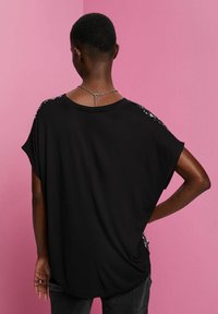 Svart kortärmad topp med lös passform, med paljettdetaljer på axlarna. Slät textur, rund halsringning och design med hög-låg fåll.