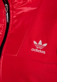 Červená bunda s lesklou, hladkou texturou a matnou fleecovou částí. Bílé logo Adidas se třemi pruhy na hrudi, zip na přední straně.