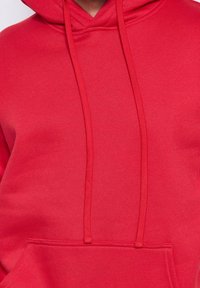 Haut à capuche rouge avec cordons et poche kangourou, montrant la poitrine et la partie supérieure du torse.