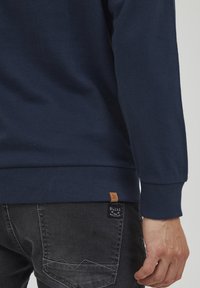 Marineblaues Baumwoll-Sweatshirt mit gerippten Bündchen und Saum. Es hat ein kleines braunes Markenetikett auf der linken Seite und graue Jeans.