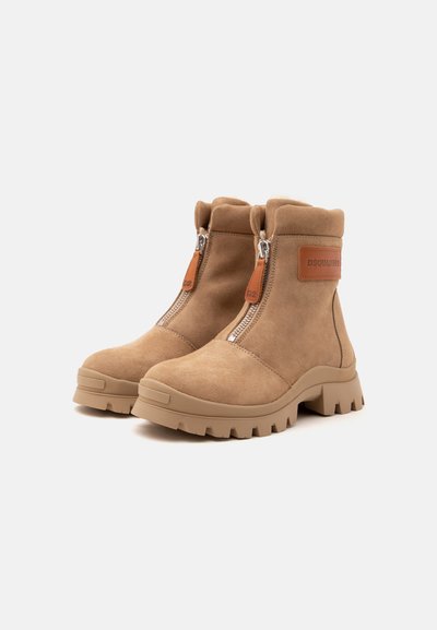 Paire de bottines en daim beige avec semelles épaisses, fermetures éclair à l'avant avec languettes en cuir, et un écusson logo sur le côté.