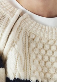 Maglione crema con texture e motivi a treccia e pompon. Presenta una zip argentata e strisce a contrasto di colore blu navy all'orlo.