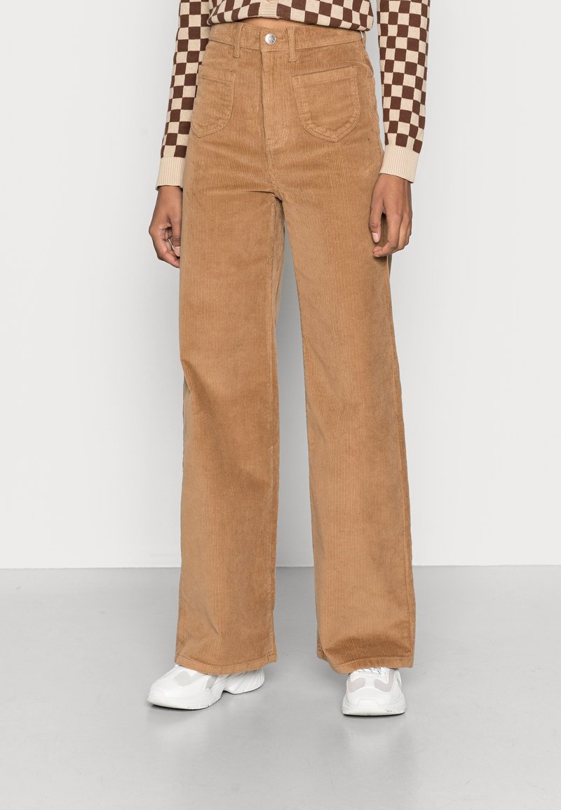 Personne portant un pantalon large taille haute en velours côtelé marron, des baskets blanches et un haut à manches longues marron et beige à carreaux.