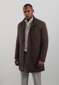 Hnědý kabát s vysokým límcem, na zip a se všitoumi kapsami. Nositelný přes světle šedý svetr a kalhoty, s hladkou texturou.