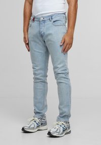 Férfi világoskék, slim fit farmernadrágban, fehér ujjatlan felsőben és ezüst színű sportcipőben áll egy sima szürke háttér előtt.