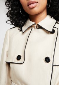 Beżowy trencz z czarną lamówką, dużymi czarnymi guzikami i kołnierzem. Posiada złote zapięcie przy szyi i gładką fakturę materiału.