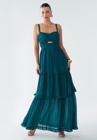 Abito lungo turchese con corpetto aderente, design incrociato sul davanti e apertura a occhiello. Tre balze a più livelli sull'orlo aggiungono texture e movimento.