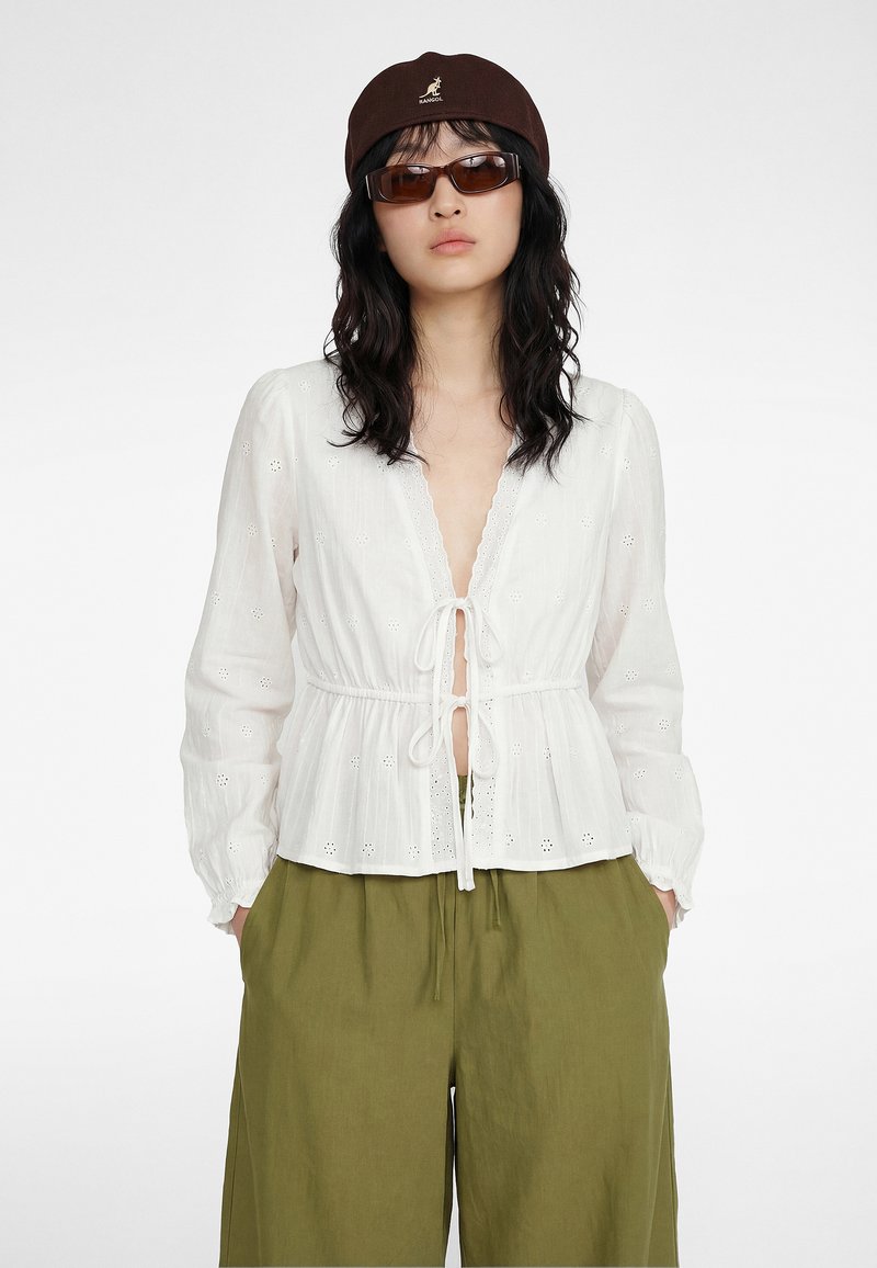 Kobieta w brązowej czapce, prostokątnych okularach przeciwsłonecznych, białej bluzce z długim rękawem z wiązaniami z przodu i luźnych oliwkowozielonych spodniach, z rękami w kieszeniach.