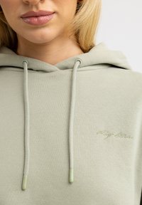 Hellgrüner Hoodie aus Baumwolle mit weicher Textur. Verfügt über ein Logo in passendem Garn und Kordeln mit klaren Kunststoffspitzen.