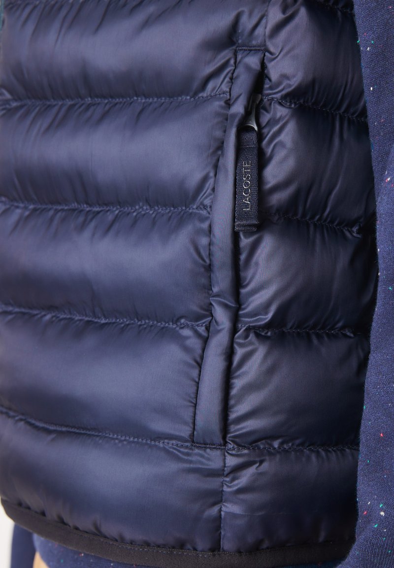 Gilet matelassé bleu marine avec un design en losanges, comportant une poche zippée sur le côté et une étiquette avec le logo Lacoste. Texture de tissu lisse et brillant.