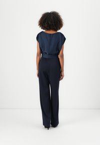 Tuta blu con top drappeggiato, maniche a cappuccio e vita evidenziata da una cintura. Pantaloni a gamba larga che arrivano fino alla caviglia. Tessuto morbido con una leggera lucentezza.