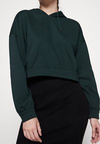 Sudadera corta de color verde oscuro con capucha ajustable, mangas largas abullonadas y puños acanalados, combinada con una falda negra acanalada.