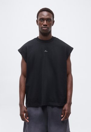 Homme aux cheveux courts portant un débardeur noir avec un petit logo blanc au centre, debout devant un fond clair uni.