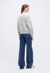 Femme aux cheveux bruns portant un sweat-shirt gris clair, un jean bleu ample et des baskets blanches, debout de côté contre un fond blanc uni.