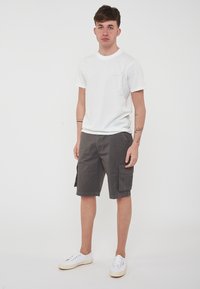 T-shirt bianco in cotone con logo, pantaloni cargo grigi con tasche laterali e sneakers bianche, in piedi su uno sfondo chiaro.