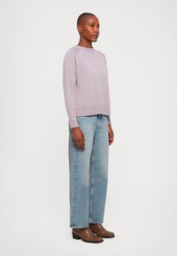 Woman wearing a light purple sweater, light blue wide-leg jeans, and brown ankle boots, standing against a plain white background.