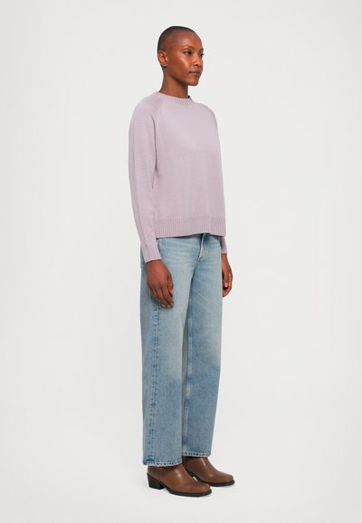 Mulher vestindo um suéter lilás claro, jeans de perna larga azul claro e botas castanhas até o tornozelo, posando contra um fundo branco simples.