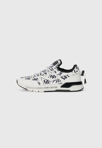 Hvit sneakers med svart mønstret tekst, har en strukturert tekstiloverdel, kontrasterende svarte detaljer, og har en merkevaret såle og hæltab.