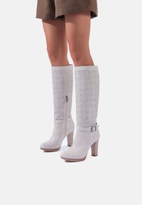 Stivali bianchi in pelle al ginocchio con design perforato, tacchi robusti, zip laterale e dettaglio della fibbia. Finitura morbida e forma strutturata.