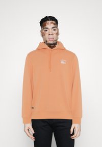 Un sweat à capuche orange en tissu doux, doté d'une poche avant, d'un cordon réglable et d'un logo Lacoste brodé sur la poitrine.