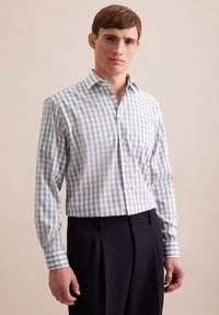 Camicia a quadri blu e bianca con maniche lunghe, design con colletto e bottoni bianchi, indossata con pantaloni scuri.