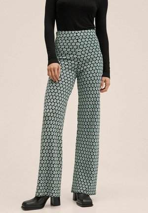 Personne portant un pantalon taille haute évasé à motif fleuri bleu clair, un haut noir à manches longues et des chaussures compensées noires.
