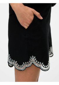 Zwarte rok met een gegolfde zoom, voorzien van witte geborduurde accenten en delicate uitsnijdingen. Het materiaal lijkt lichtgewicht en glad te zijn.