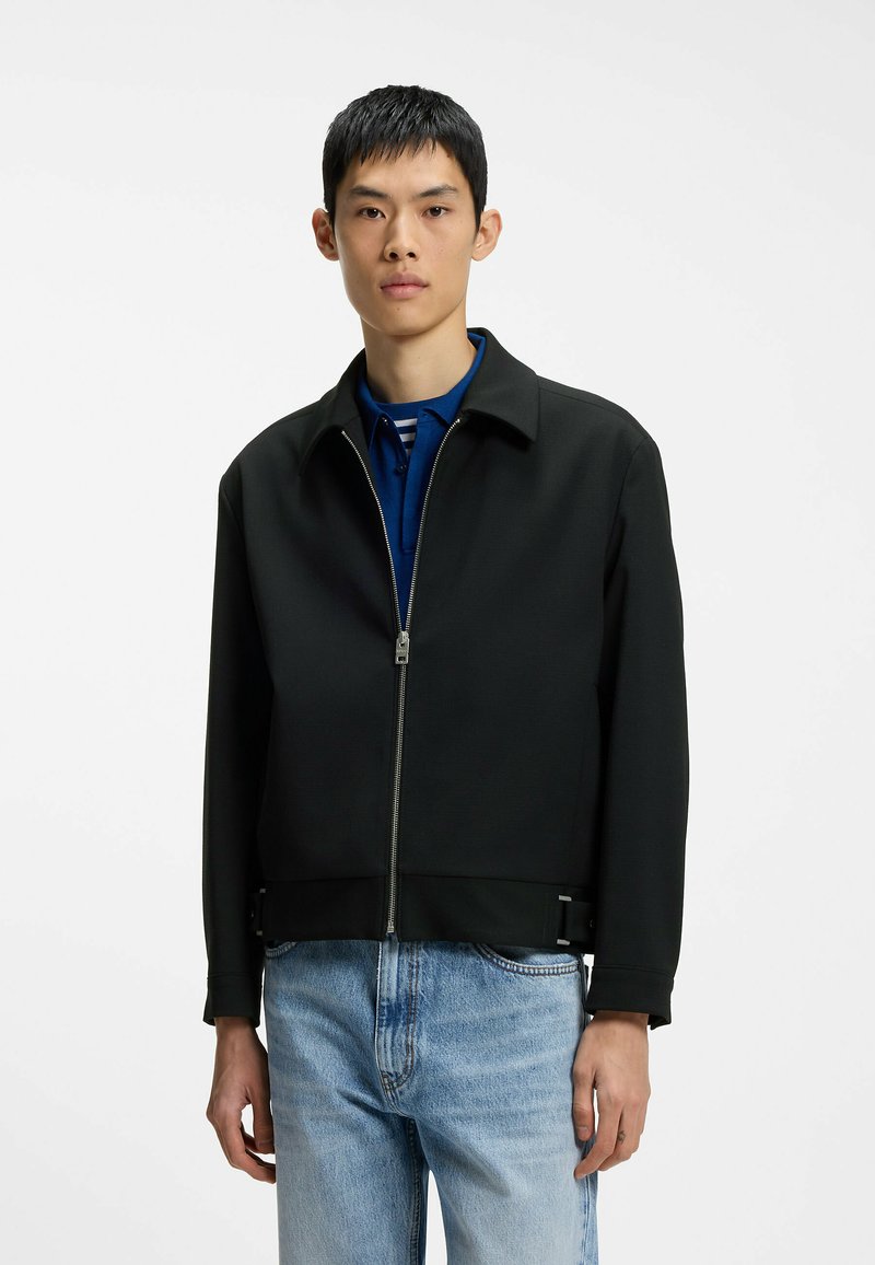 Jeune homme portant une veste zippée noire sur une chemise bleue à col et un jean bleu clair, debout devant un fond blanc uni.